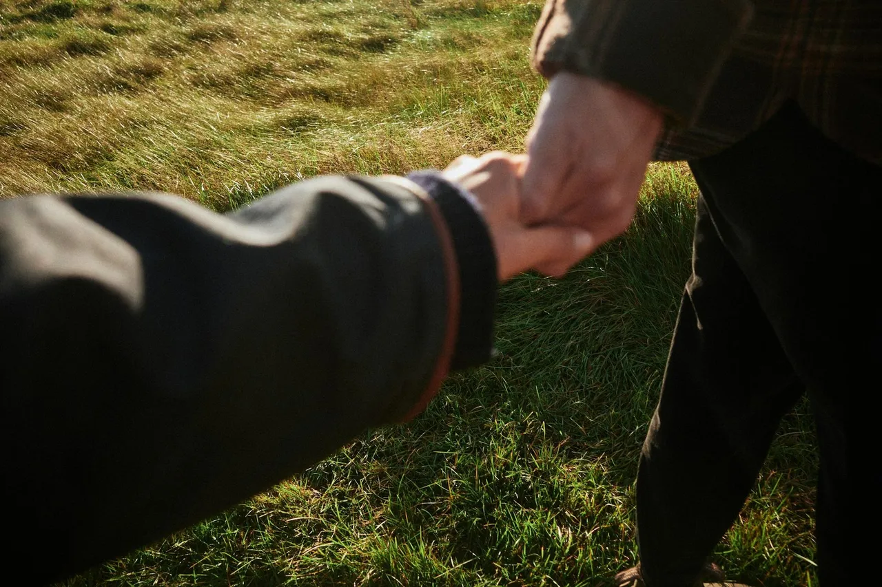 Two people holding hands over a sunlit field of grass.